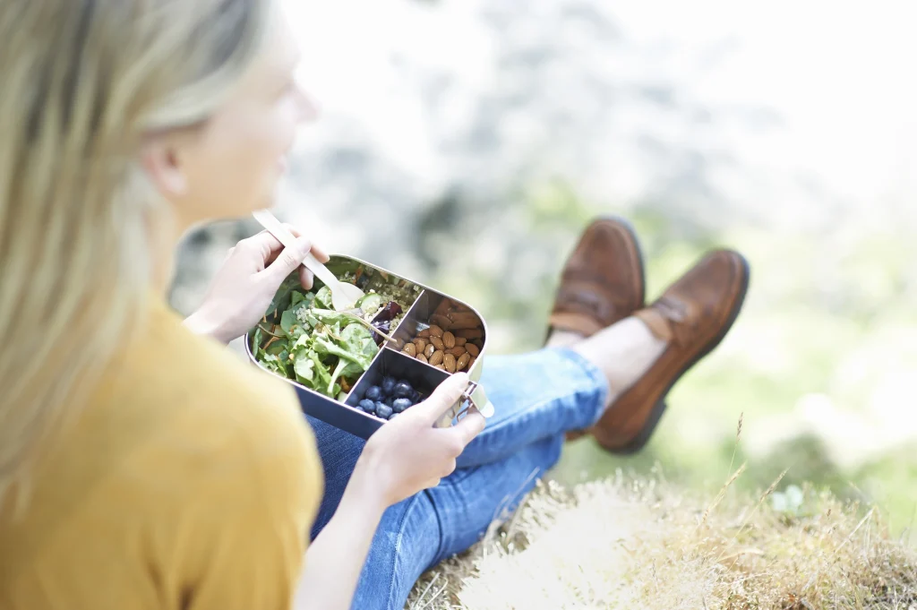woman sits eating a healthy lunch of fruit, nuts and bulgar wheat salad from a reusable plastic free lunch box in the countryside