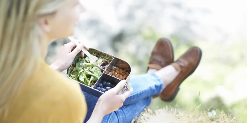 woman sits eating a healthy lunch of fruit, nuts and bulgar wheat salad from a reusable plastic free lunch box in the countryside