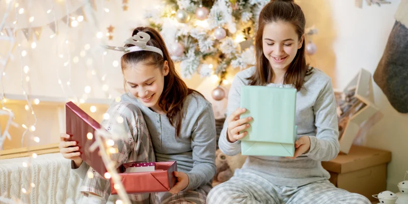 Two happy teenage girls in pajamas sitting by the Christmas tree and looking inside gift boxes