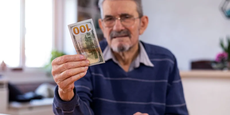 Elderly man holding a one hundred dollar bill in hopes to save it instead of spending it.