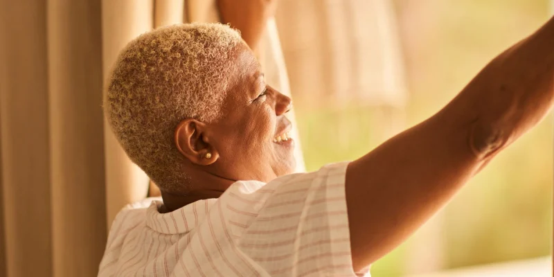 older senior woman bathed in sunlight opening a window and smiling